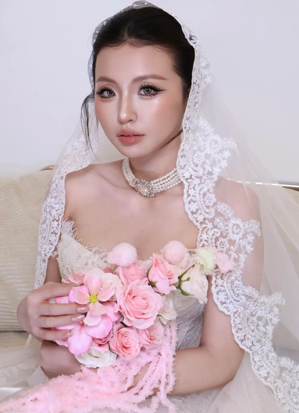 Cô dâu Yến Baby Makeup nhìn thẳng, da căng bóng, voan ren dài, vòng cổ ngọc trai nhiều tầng, ôm bó hồng phấn