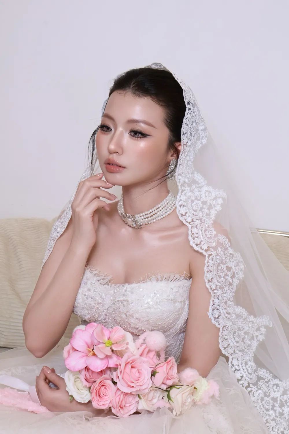 Cô dâu nghiêng mặt tay đỡ cằm, voan ren dài, bó hoa hồng phấn đặt trước mặt